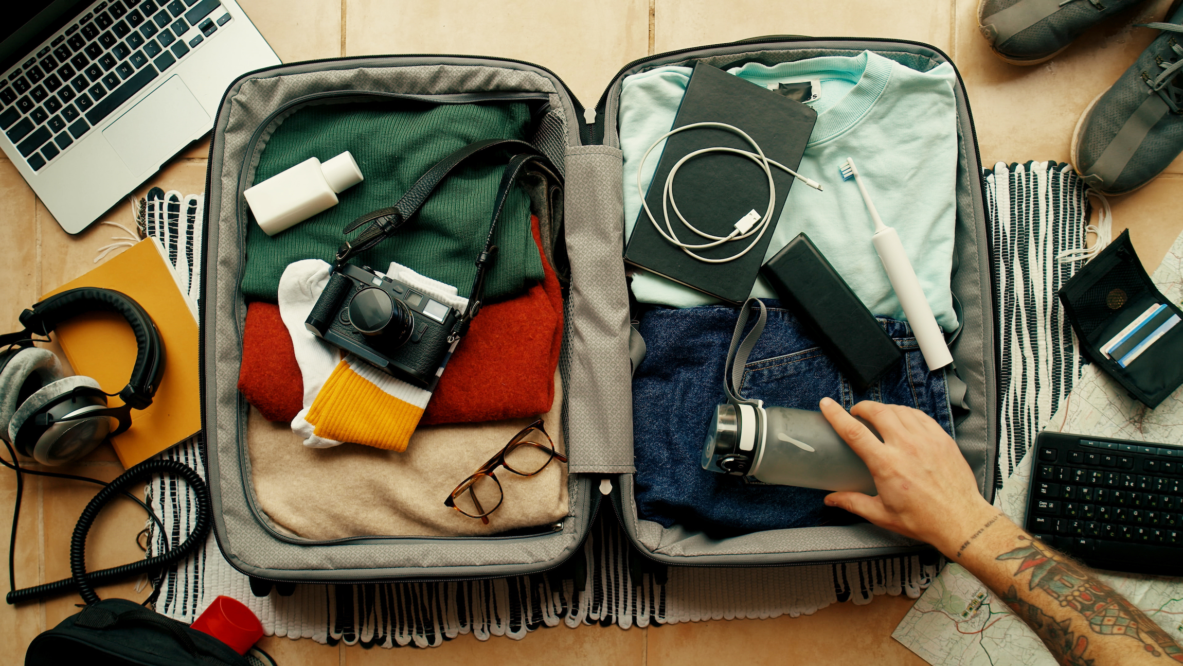 Mężczyzna z wytatuowanym ramieniem pakuje walizkę na wycieczkę. Torba z aparatem fotograficznym, laptopem, przenośną ładowarką i elektryczną szczoteczką do zębów. Nowoczesna koncepcja blogera podróżniczego